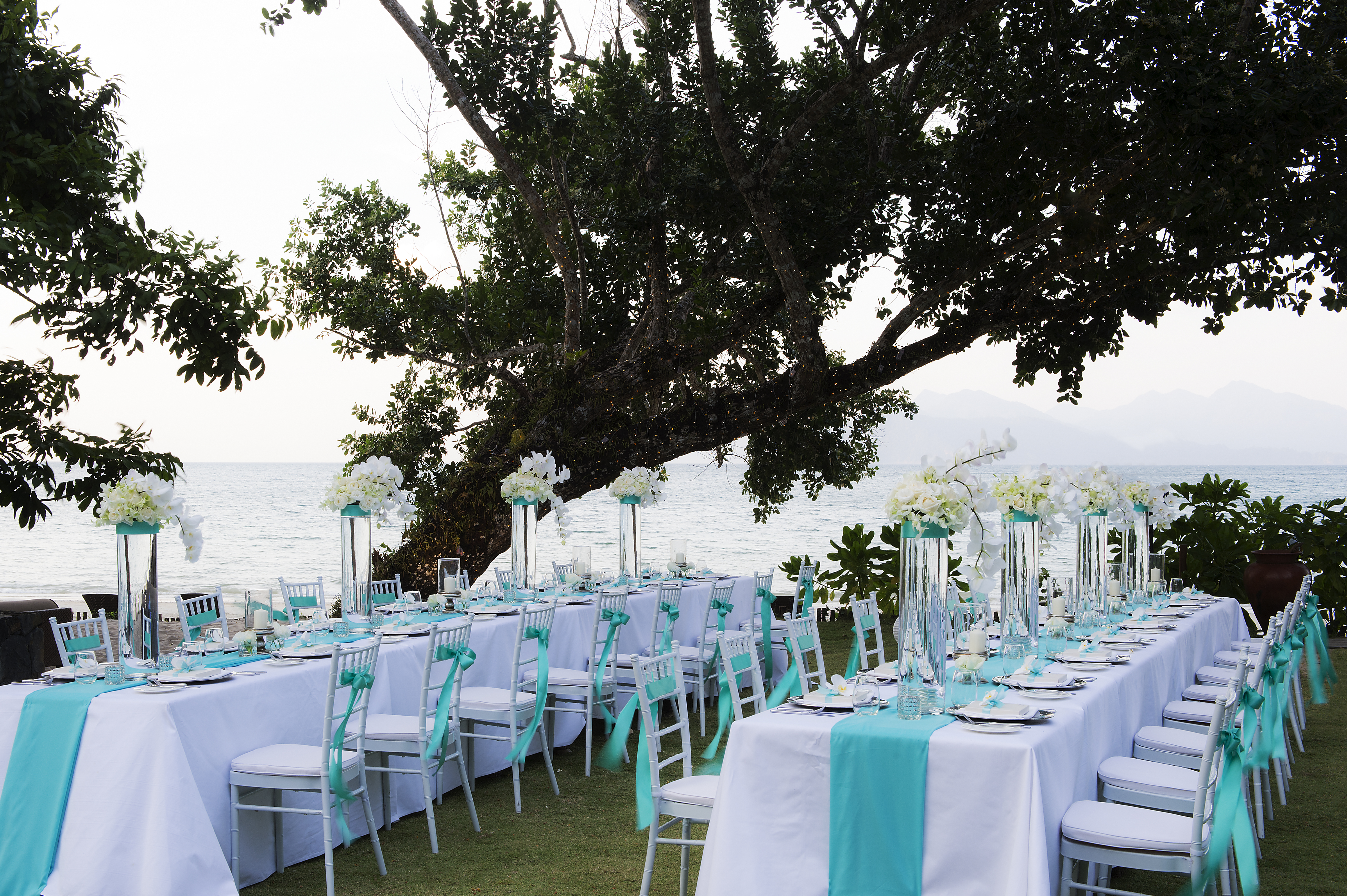 Two long blue and white tables equipped with chairs  and decorated with flowers in tall glasses overlooking the datai bay 